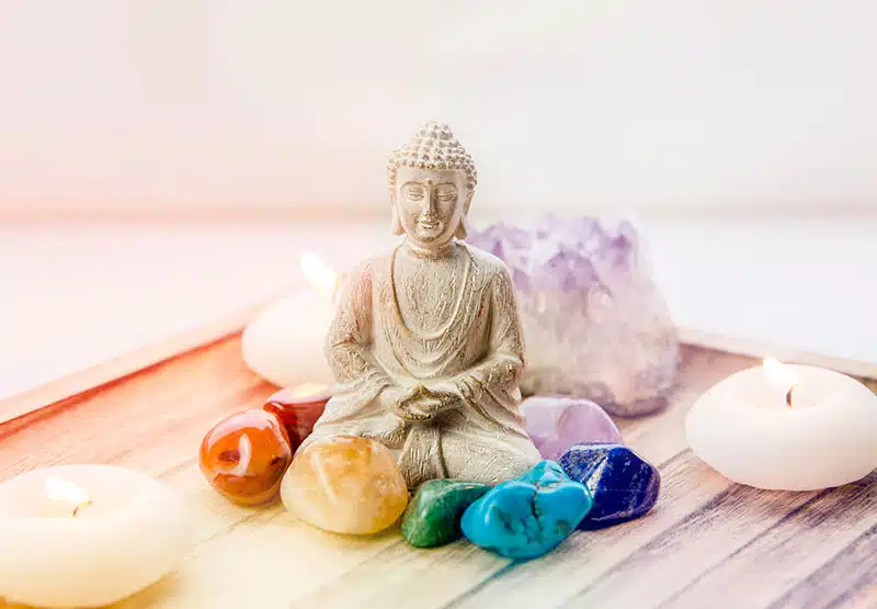 Meditation-800 Buddhafigur, klein, weiß, umgeben von bunten Edelsteinen und zwei kleinen, runden Kerze auf einem Tablett