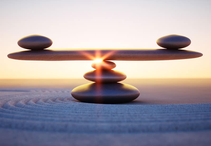 Steine an einem Stand, die zu drit zu einem kleinen Turm gelegt sind, auf dem ein großer flacher Stein ruht. Auf diesem ruhen an jedem Ende ein weiterer, flacher Stein als Symbol der Balance.