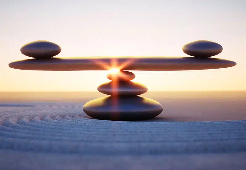 Steine an einem Stand, die zu drit zu einem kleinen Turm gelegt sind, auf dem ein großer flacher Stein ruht. Auf diesem ruhen an jedem Ende ein weiterer, flacher Stein als Symbol der Balance.